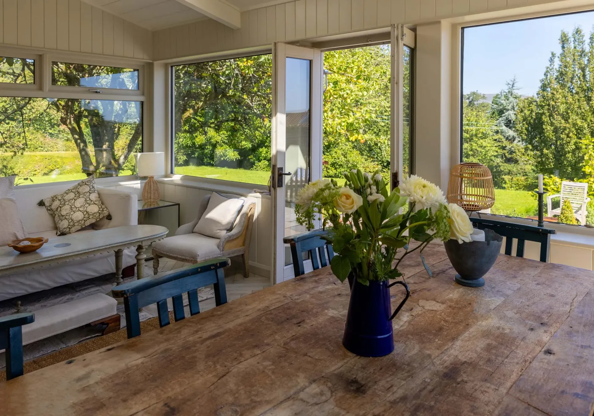 Sunroom interior with wooden table, floral arrangement, and view of green trees.
