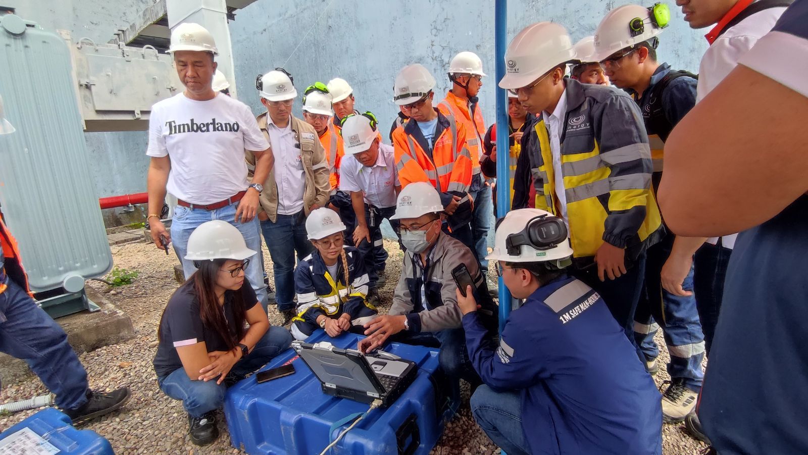 A group of people wearing hard hats and safety vests are standing around a blue box.