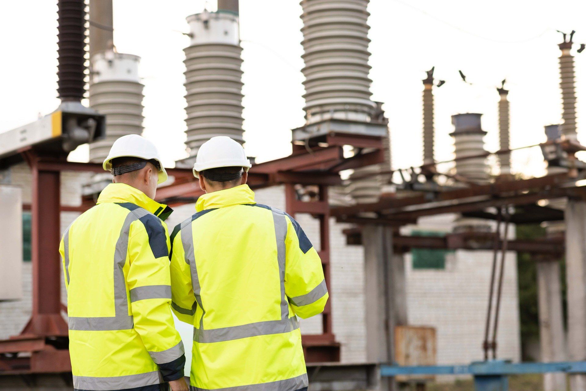 Two men in yellow safety jackets and hard hats are standing in front of a power station.