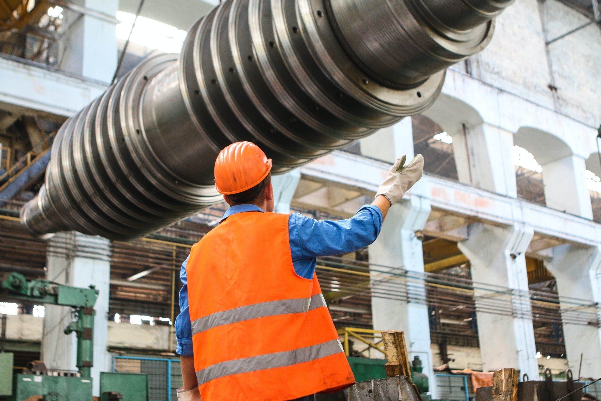 A man in an orange vest and hard hat is standing in front of a large pipe in a factory.