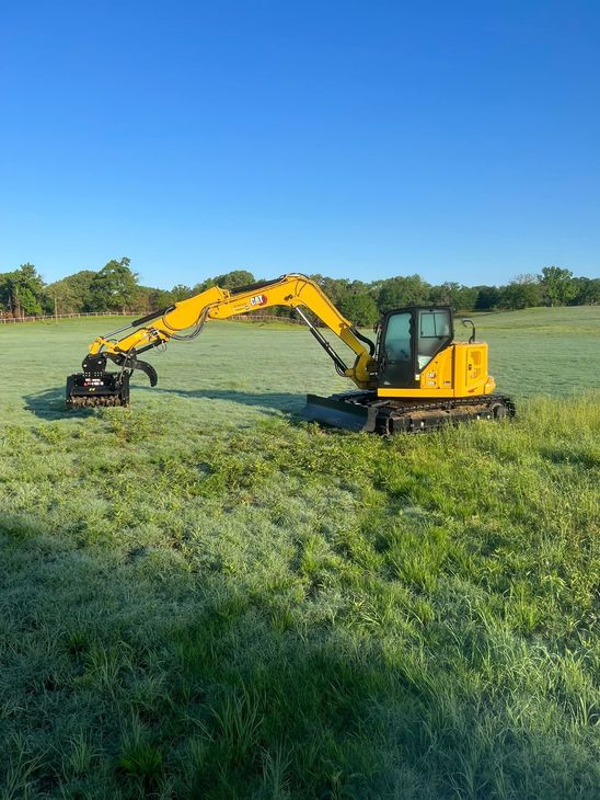 Yellow excavator on green grass under a blue sky, operating a tree shear attachment.