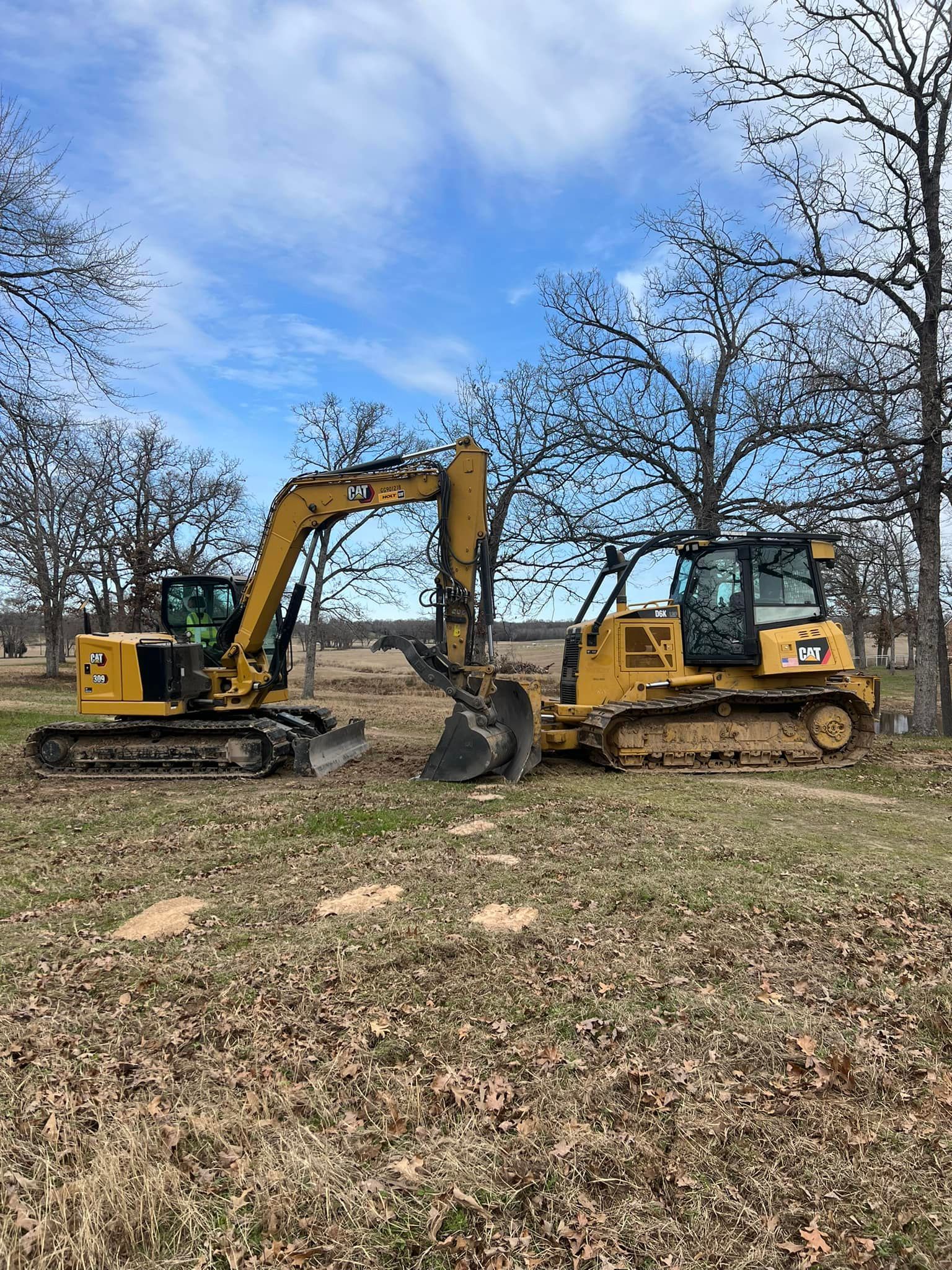 Yellow excavator and bulldozer on grassy land, blue sky with trees in background.