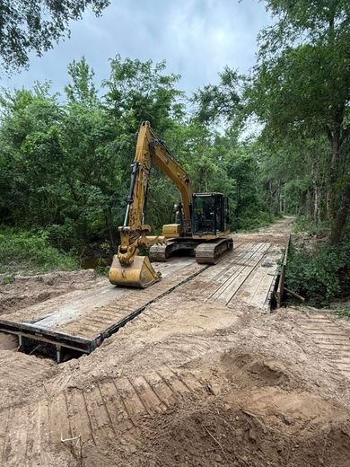 Yellow excavator on a wooden bridge in a wooded area, likely for construction or repair.