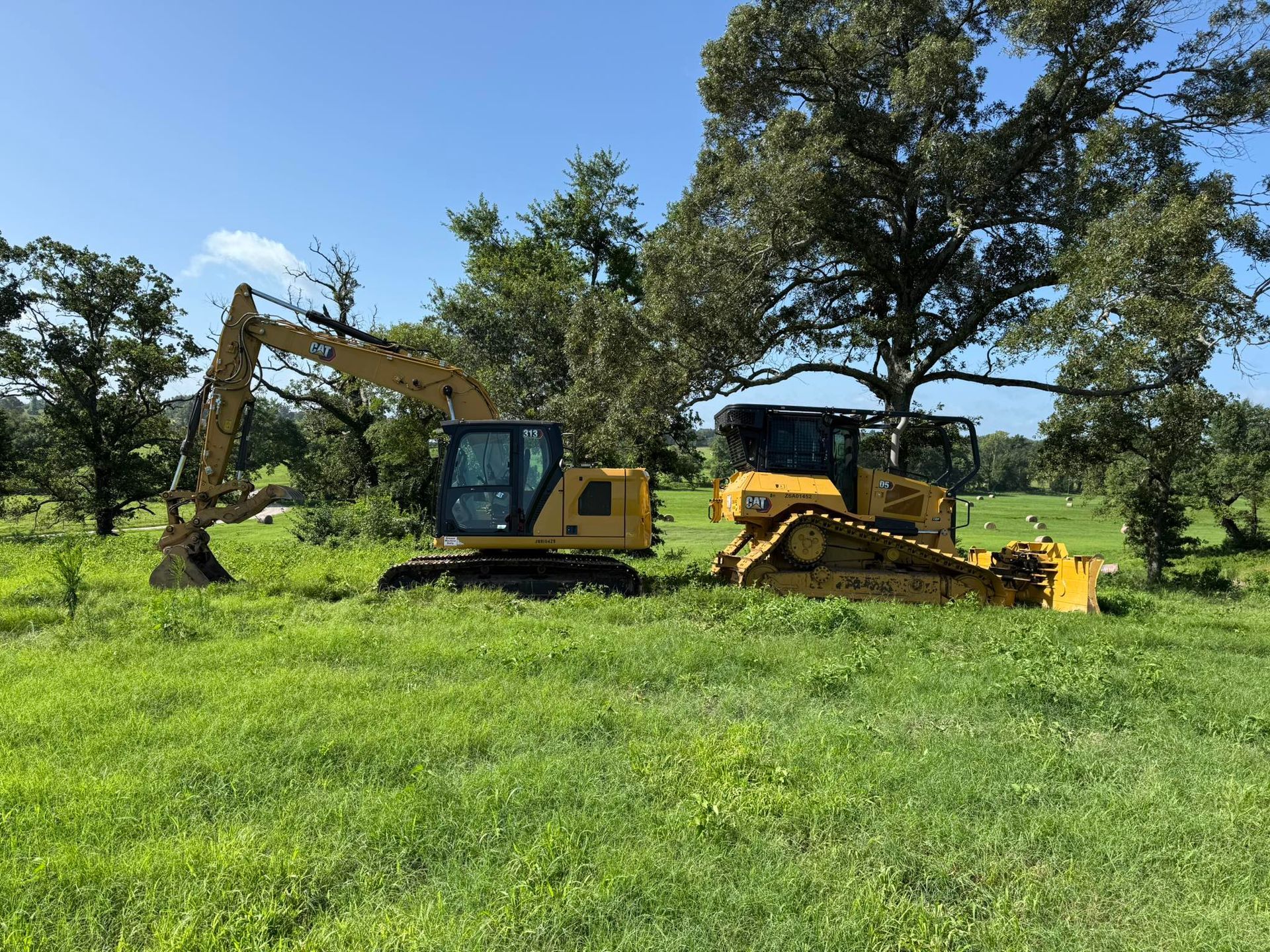 Yellow excavator and bulldozer in a grassy field with trees under a blue sky.
