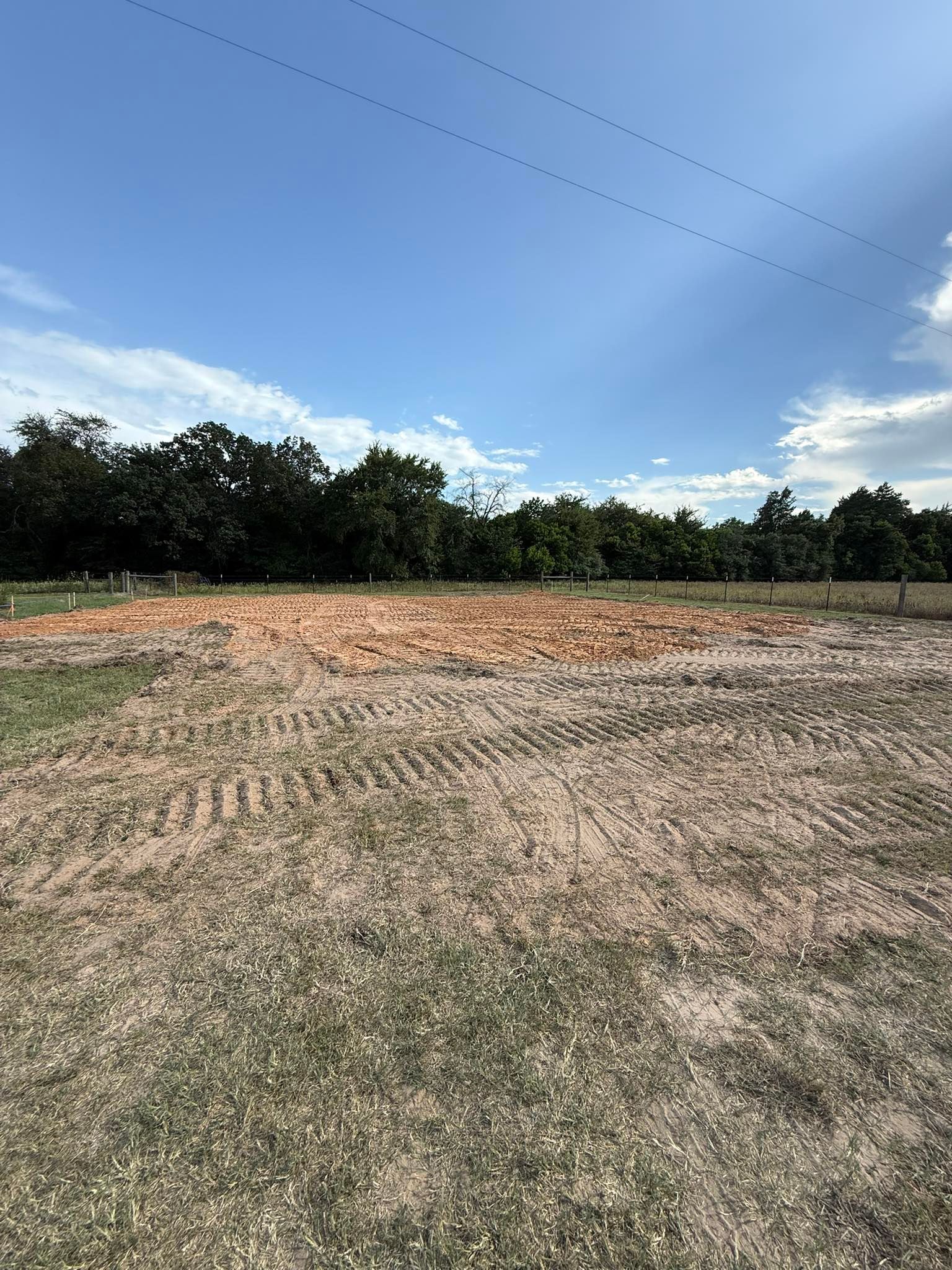 Dirt field, brown and gray, with green grass in the foreground under a blue sky. Trees in the background.