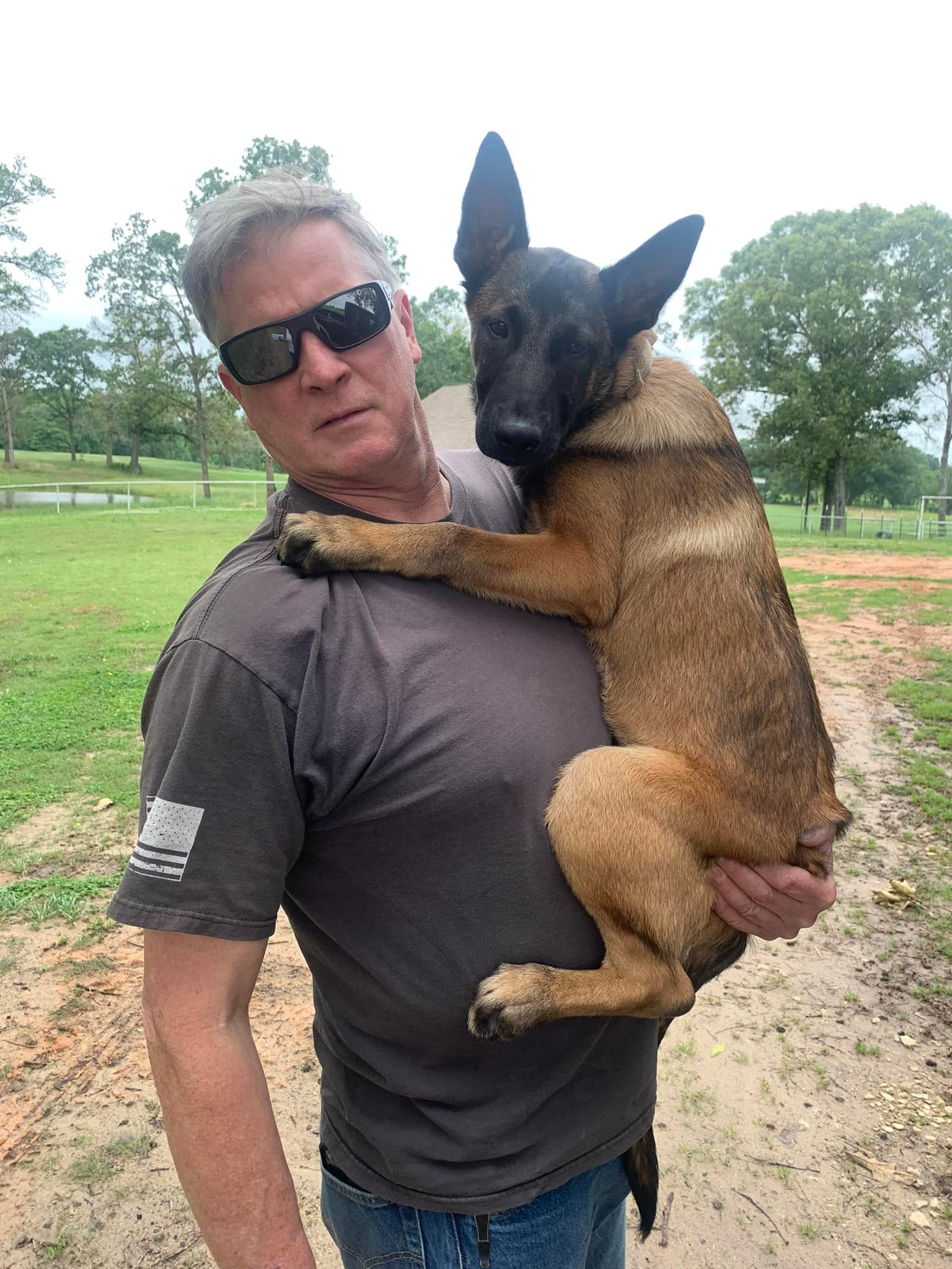 Man in sunglasses carrying a brown dog over his shoulder on a path in a grassy field.