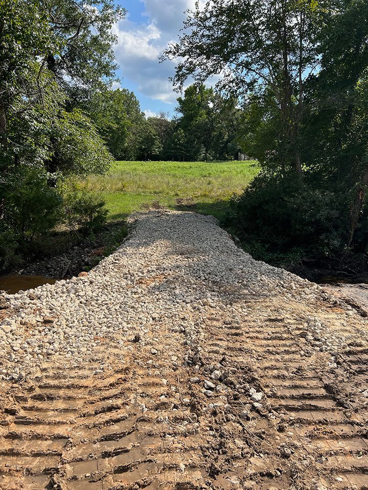 Gravel road crosses a shallow creek.  Tire tracks in dirt lead to the gravel.  Green field and trees beyond.