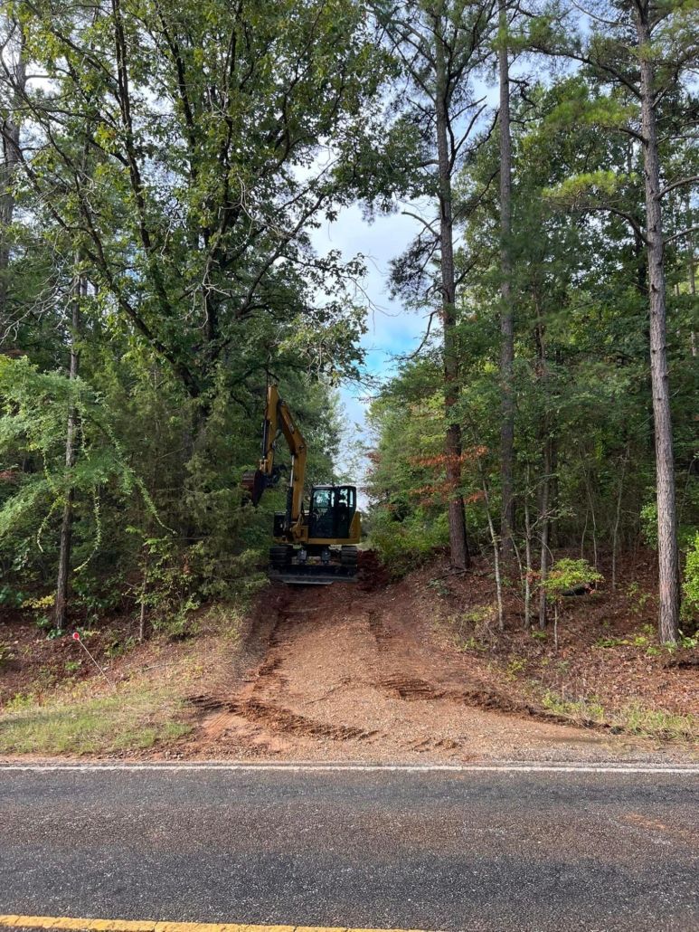 Excavator clearing brush on a wooded hillside next to a road.