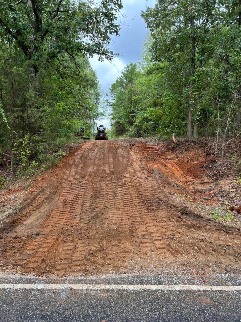 Dirt road ascends towards treeline; vehicle at top. Red-brown earth, green trees, overcast sky.