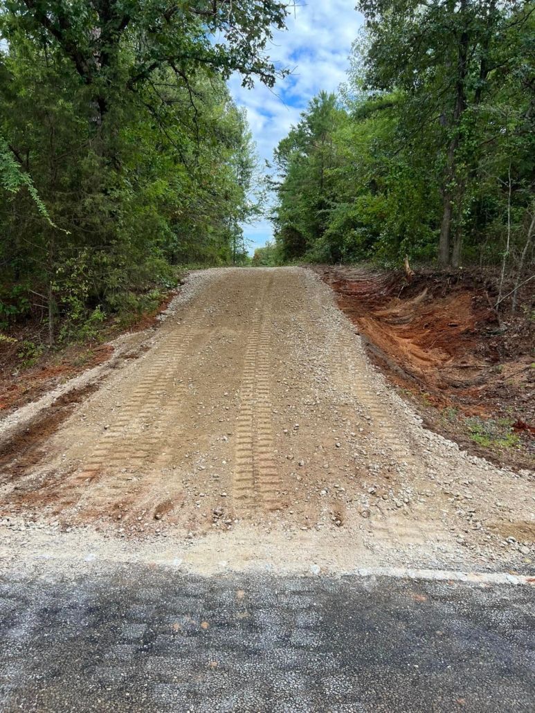A gravel road slopes upwards into lush green trees under a cloudy sky.