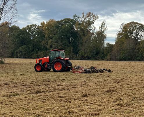 Orange tractor plowing a field; trees in the background under a cloudy sky.