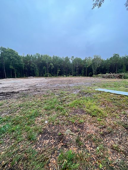 Cleared land with green grass foreground, dirt patch, and tree line under overcast sky.