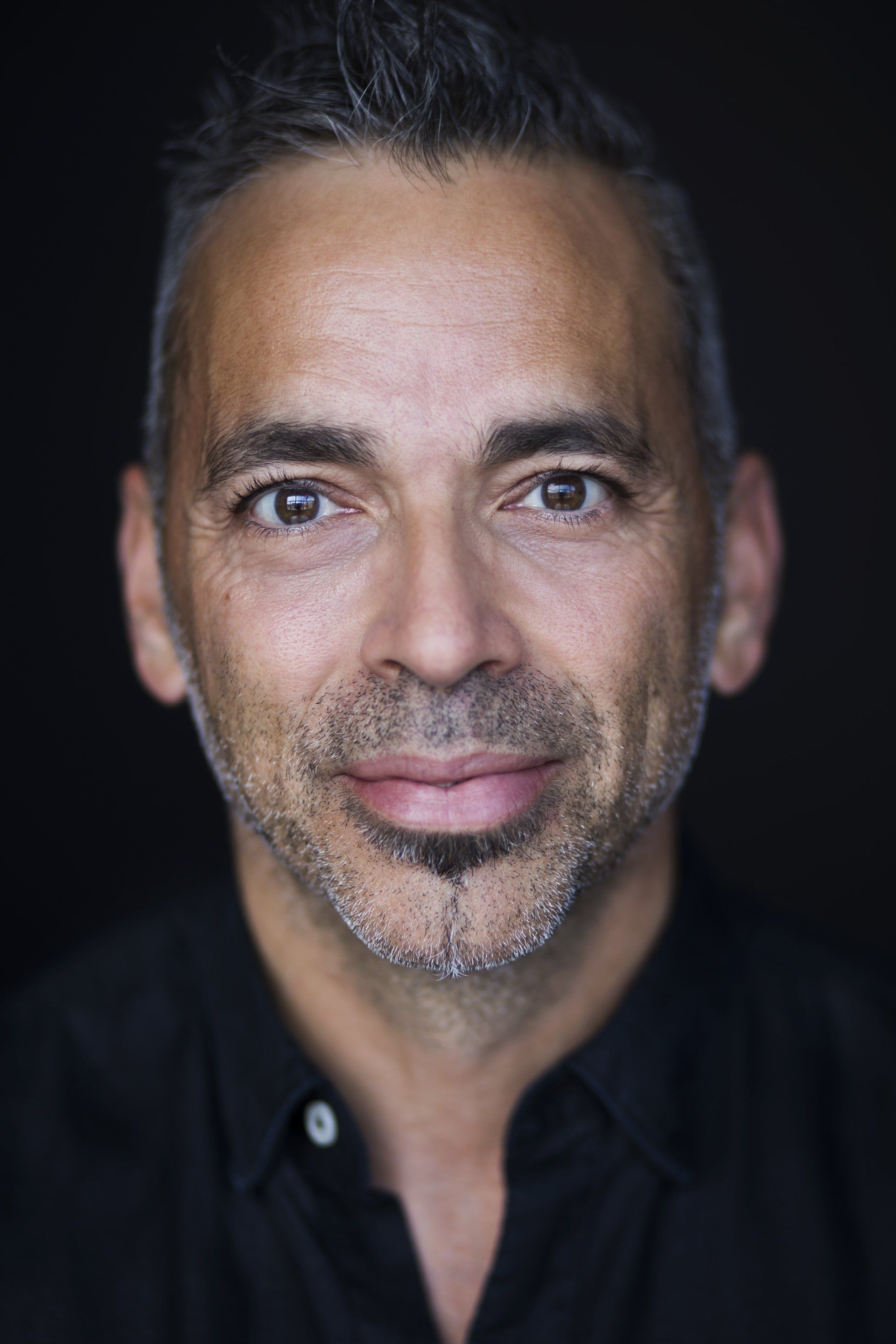Un homme aux cheveux courts et grisonnants, portant une barbe de trois jours, sourit doucement, vêtu d'une chemise noire sur fond sombre.