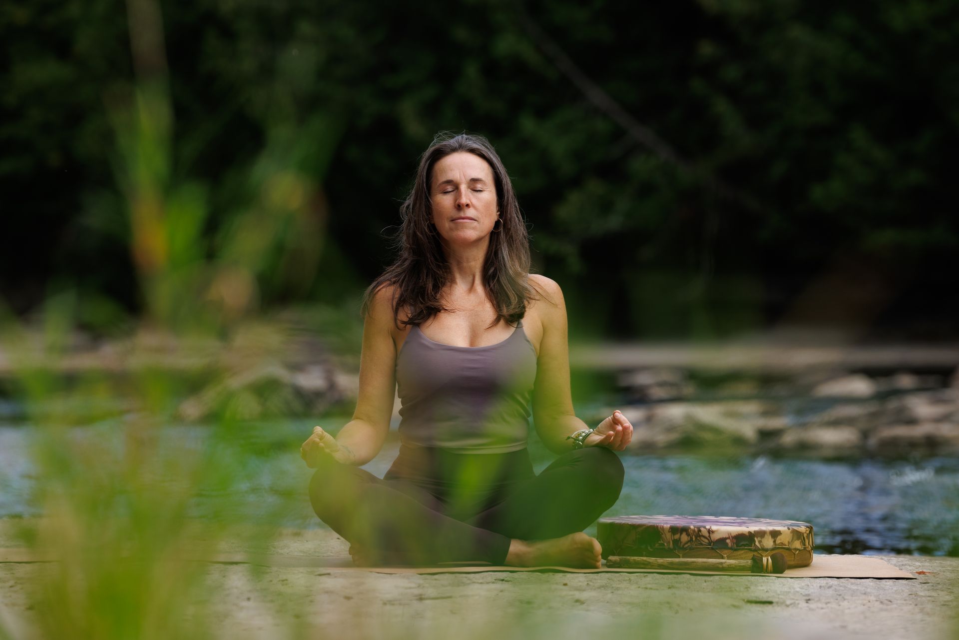 Femme méditant en plein air, yeux fermés, position du lotus, débardeur gris, pantalon vert. Arrière-plan flou de feuillage et d'eau.