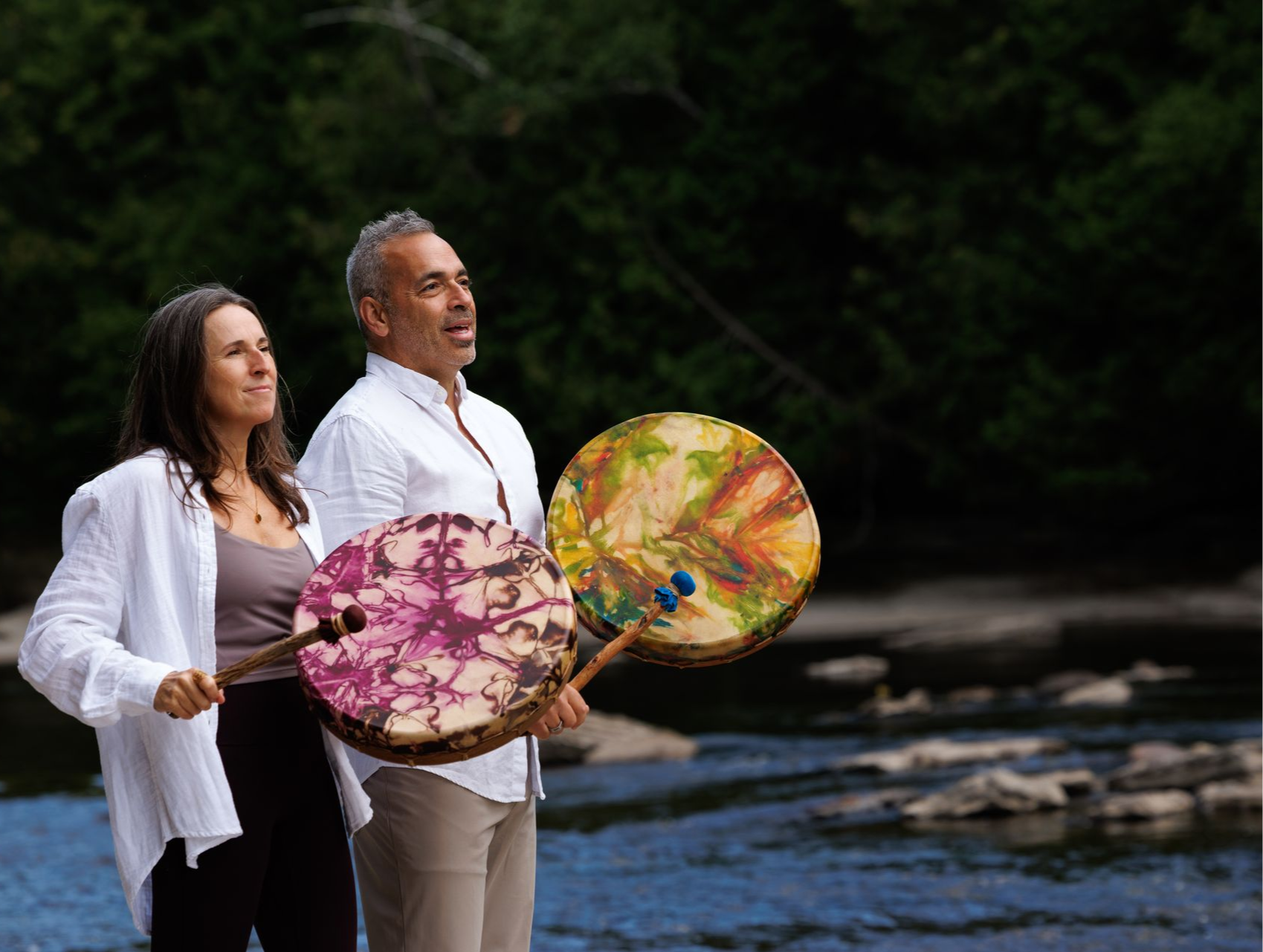 Deux personnes tenant des tambours à main près d'une rivière, le regard tourné vers le ciel. Les tambours arborent des motifs colorés.