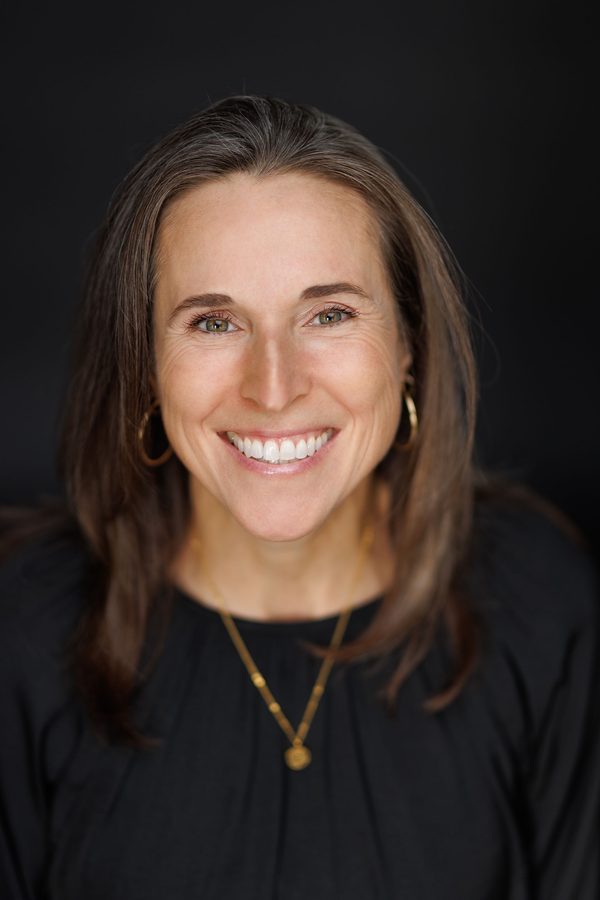Une femme sourit à l'objectif, vêtue d'un haut noir, d'un collier en or et de boucles d'oreilles créoles, sur un fond sombre.