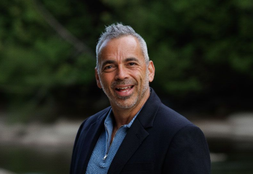 Homme aux cheveux grisonnants, souriant en extérieur, portant un blazer foncé sur une chemise bleue, sur fond vert flou.