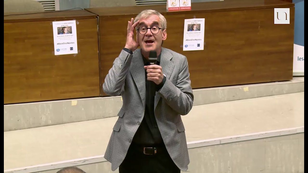 Un homme portant des lunettes fait des gestes en parlant dans un microphone lors d'une conférence.