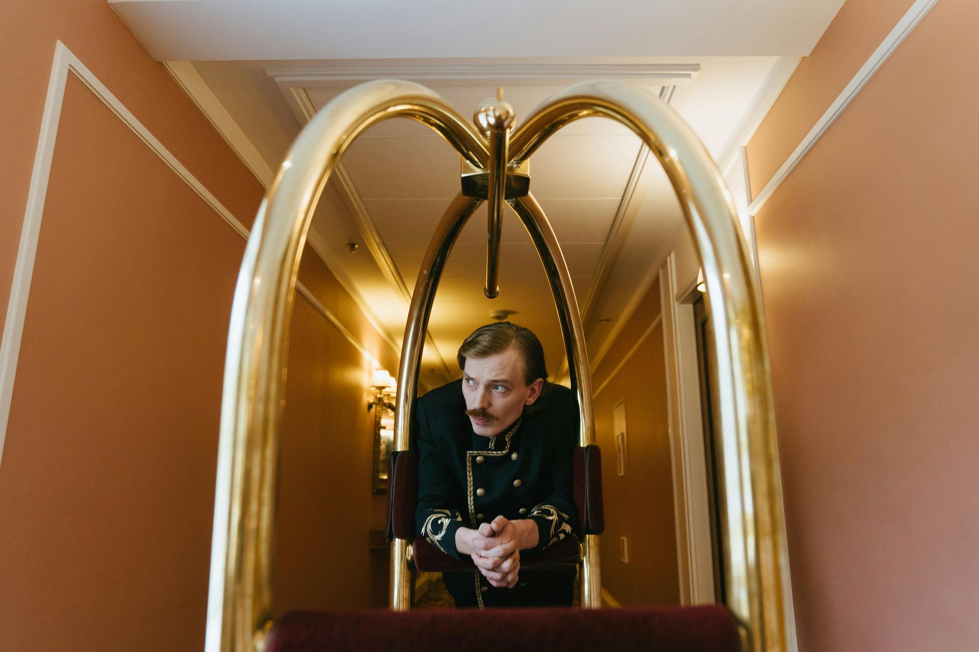 A hotel concierge in a uniform stands behind a golden luggage cart in a hallway.