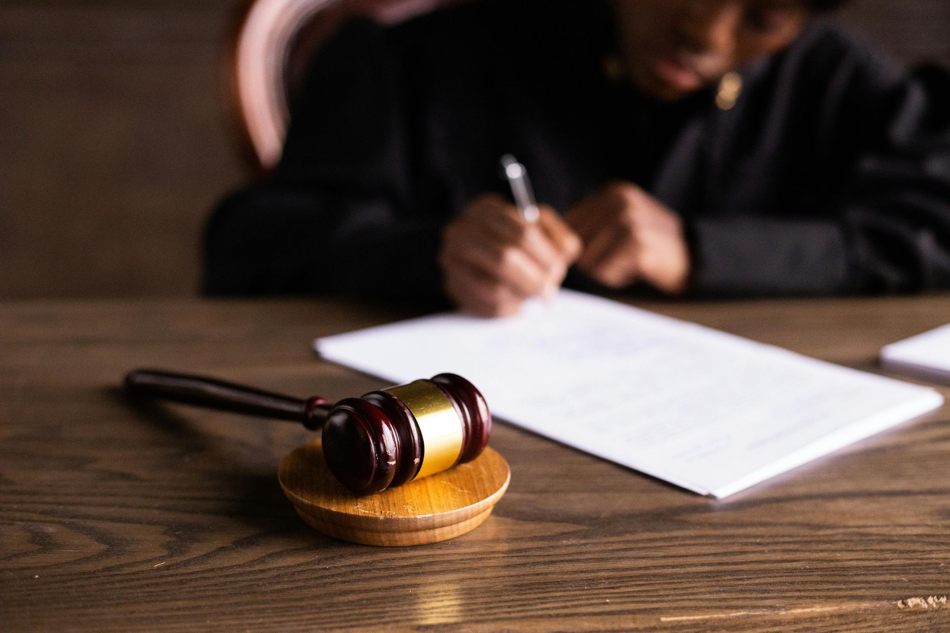 Gavel on wooden block in focus, blurred judge writing on paper in background.