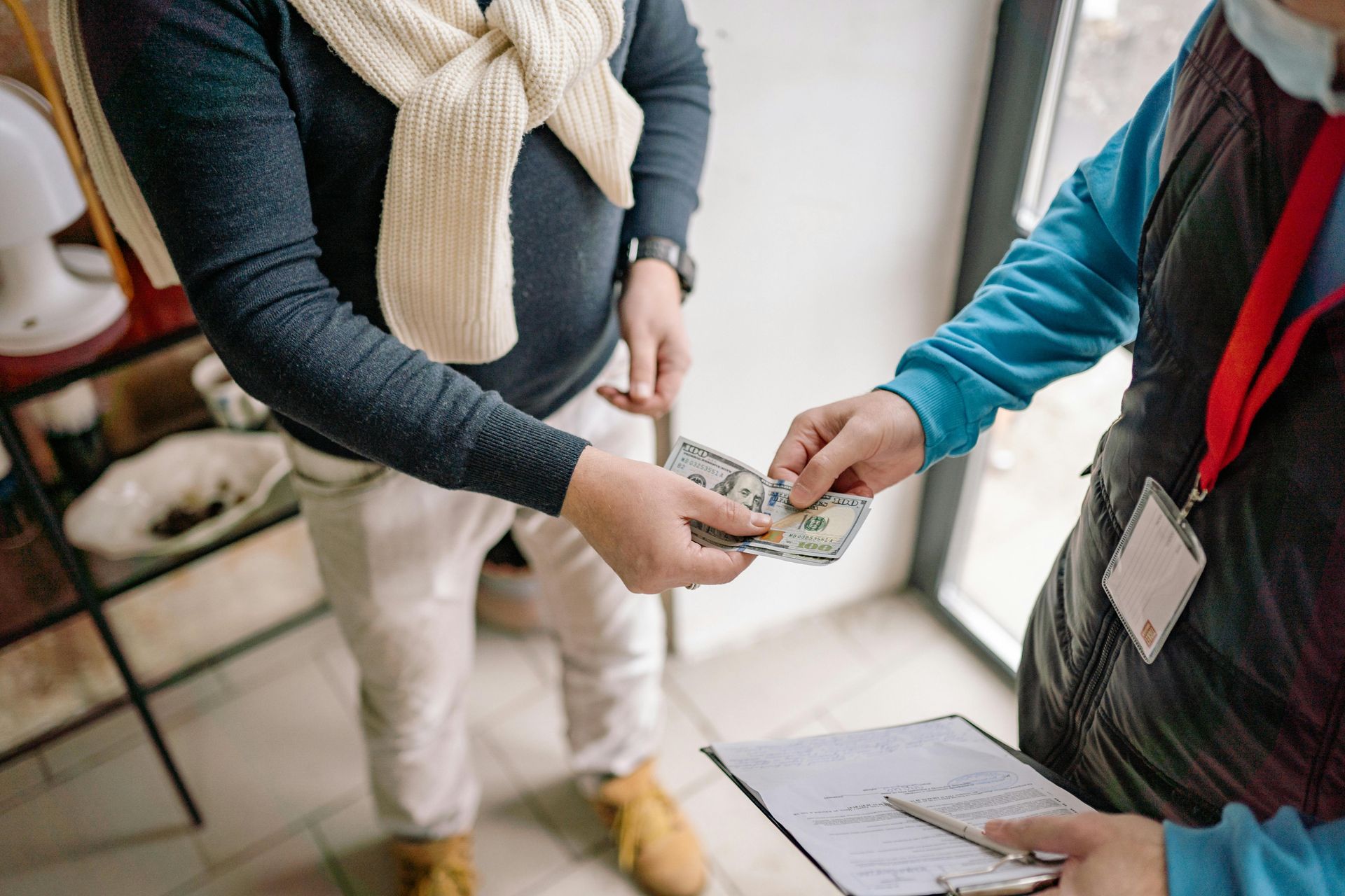 Person paying another person cash, near a window. One holds money out, other holds a clipboard.