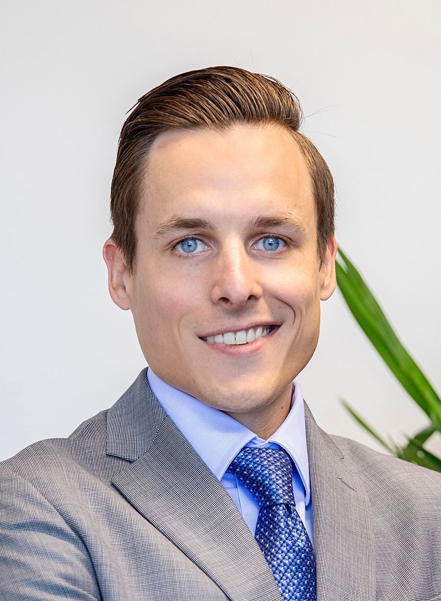 Nick Moore smiling in a gray suit and blue tie against a light background, with a green plant behind him