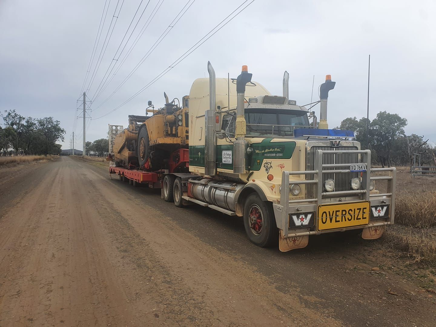 Yellow Heavy-duty Truck Hauling Machinery on a Dirt Road Under Cloudy Skies — Oakleigh Earthmoving in Chinchilla, QLD