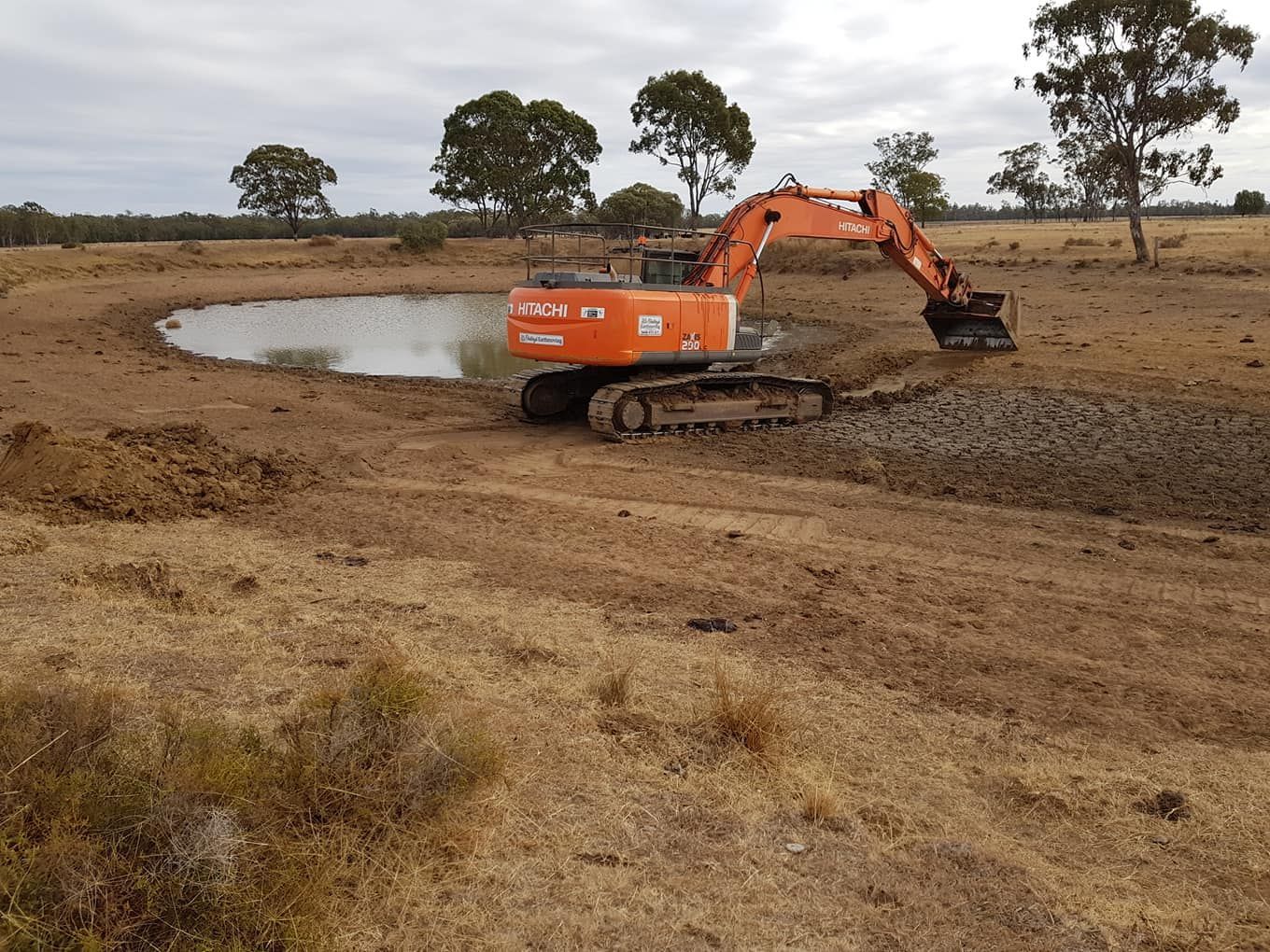 Orange Excavator Digging Beside a Muddy Pond in a Dry Field With Scattered Trees — Oakleigh Earthmoving in Kingaroy, QLD