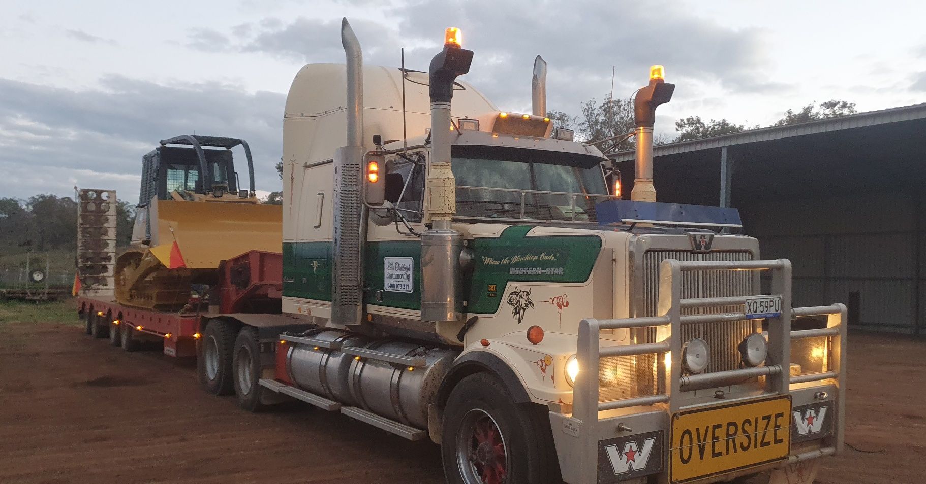 White Semi Truck Hauling a Yellow Bulldozer on a Flatbed Trailer at Dusk — Oakleigh Earthmoving in Toowoomba, QLD