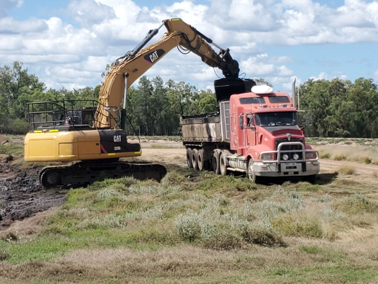 Yellow Excavator Loading Dirt Into a Red Dump Truck in a Muddy Field Under a Cloudy Sky — Oakleigh Earthmoving in Moonie, QLD