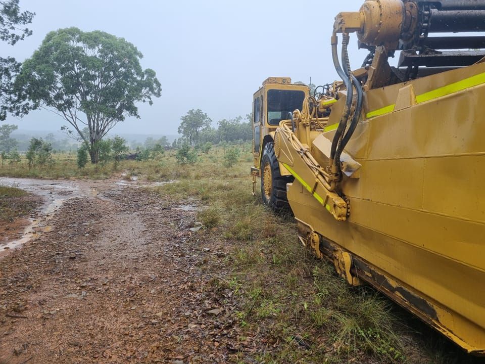 Yellow Road Grader on a Muddy Rural Dirt Road Beside Grass and Trees — Oakleigh Earthmoving in Toowoomba, QLD