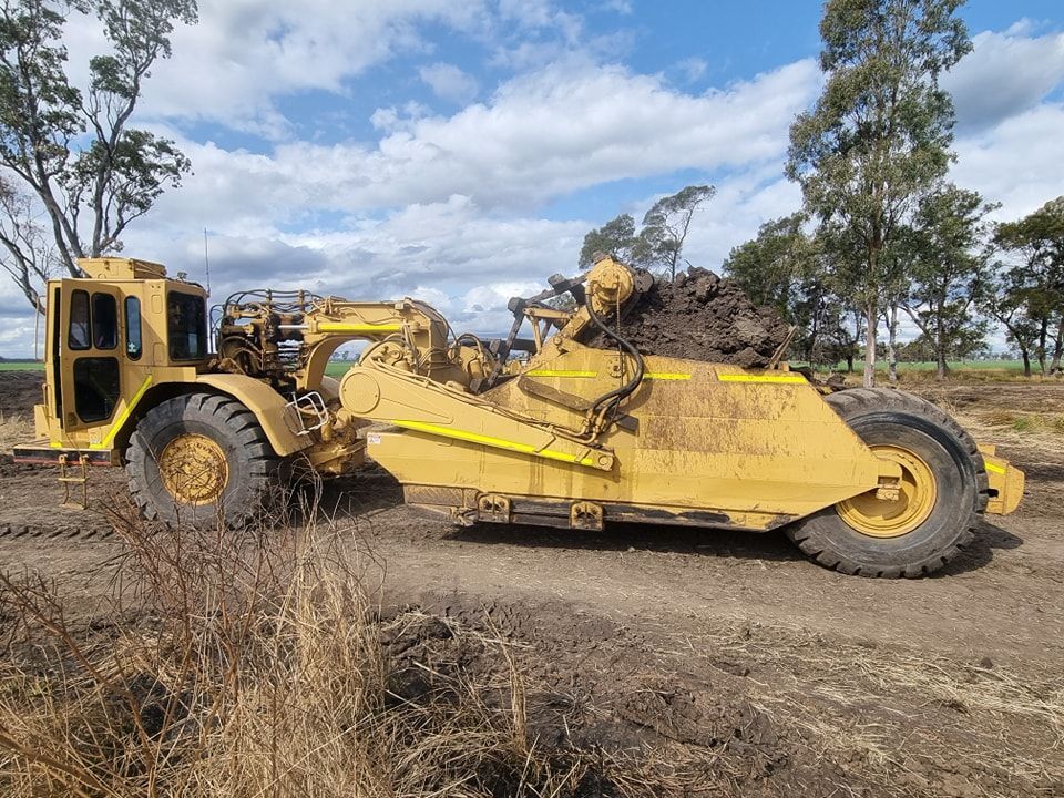 Yellow Earthmoving Scraper on a Dirt Site With Trees and Cloudy Sky — Oakleigh Earthmoving in Cotswold Hills, QLD