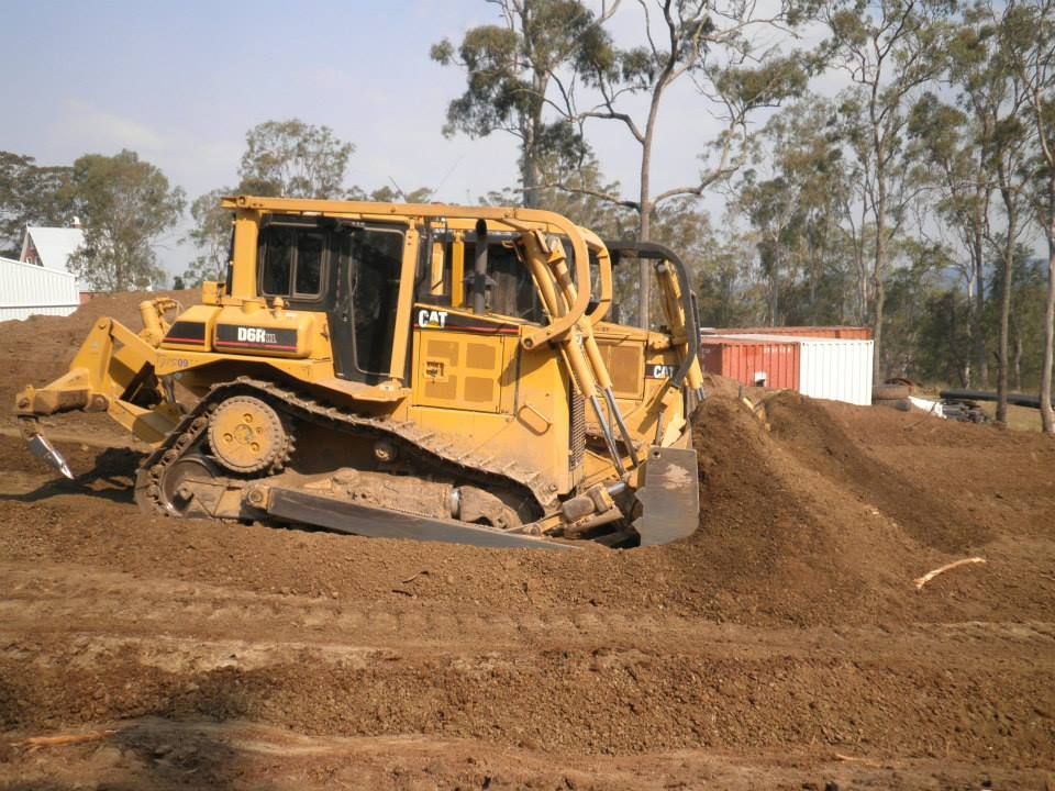 Yellow Bulldozer Pushing Dirt at a Construction Site With Trees and a Small Building — Oakleigh Earthmoving in Kingaroy, QLD