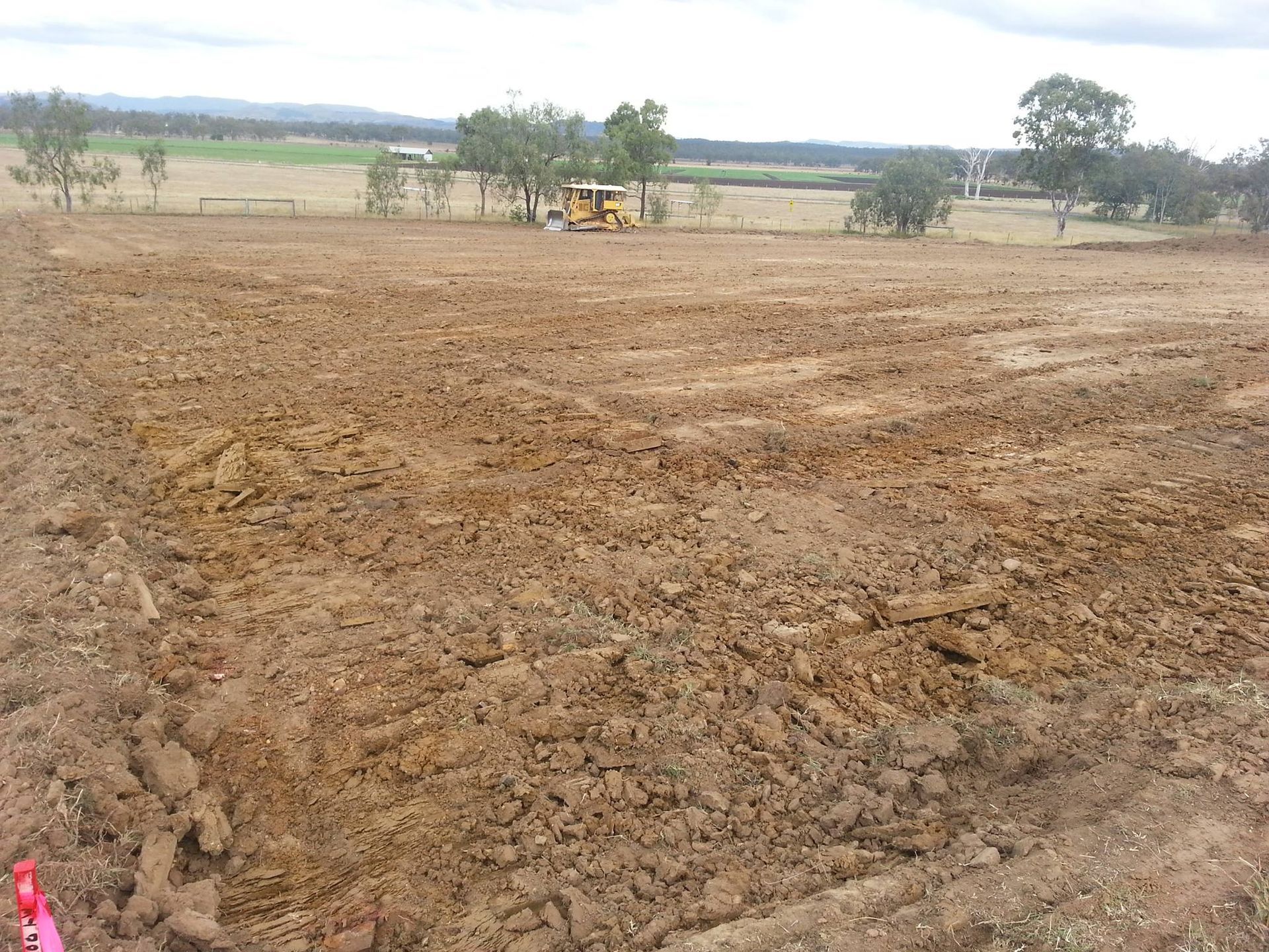 Dry Plowed Field With a Yellow Tractor in the Distance Under a Cloudy Sky — Oakleigh Earthmoving in Chinchilla, QLD