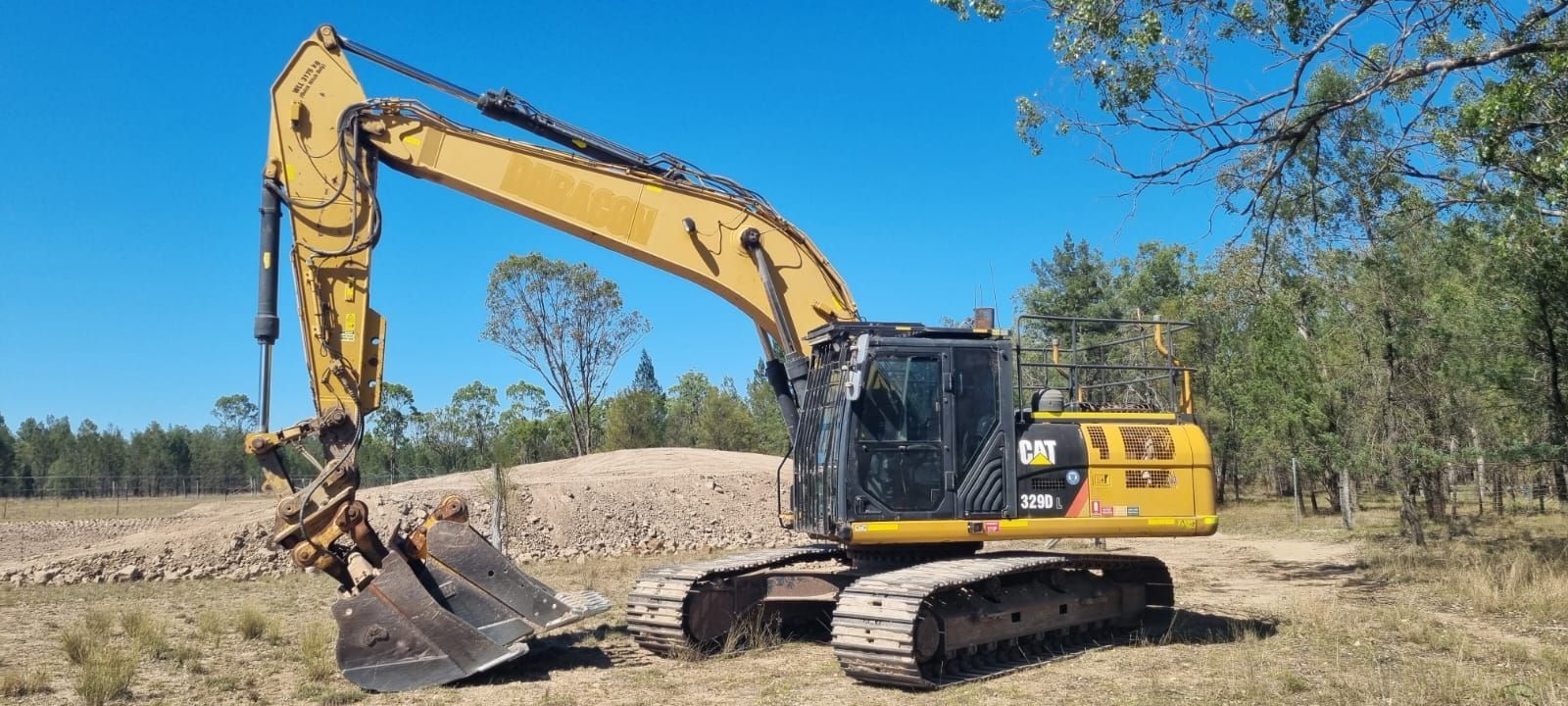 Yellow Excavator Digging on a Dirt Pile Under a Blue Sky in a Wooded Area — Oakleigh Earthmoving in Toowoomba, QLD