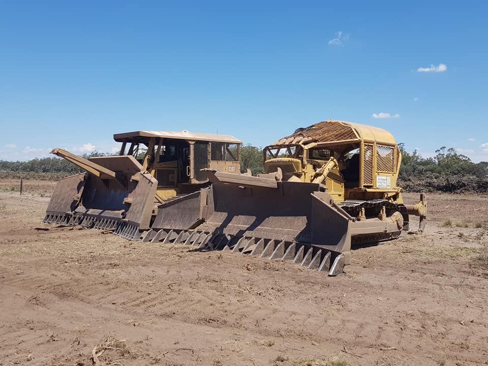 Two Yellow Bulldozers With Blades and Rakes Parked on a Dry Dirt Field — Oakleigh Earthmoving in Chinchilla, QLD
