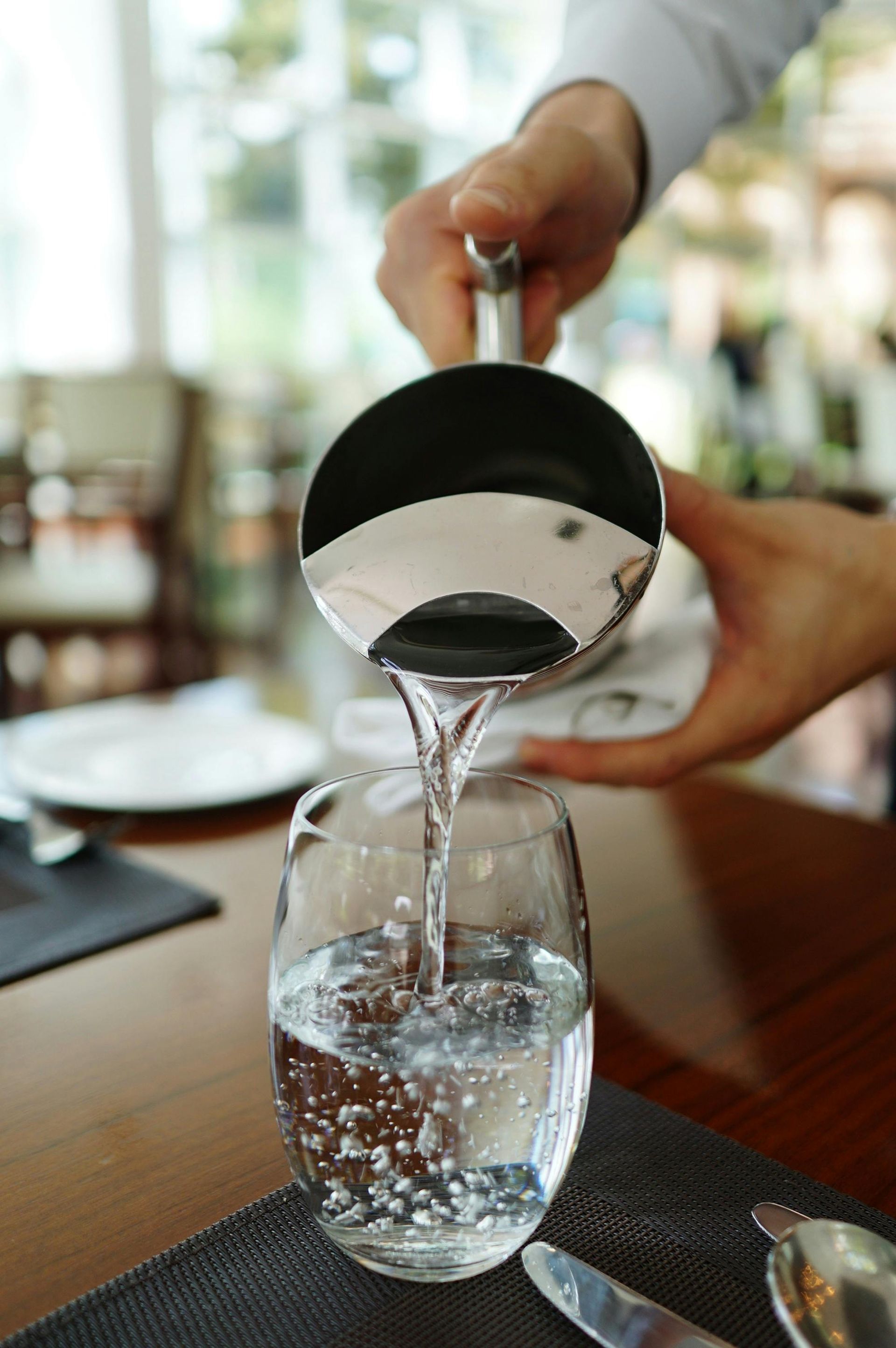 Server pouring water from a stainless steel pitcher into a glass at a restaurant table.