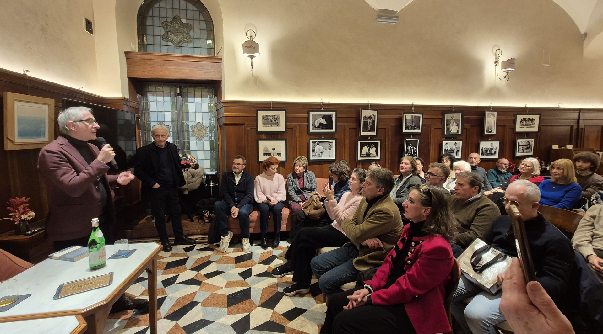 sala caffè persone sedute che ascoltano scrittore in piedi che parla con moderatore accanto che guar