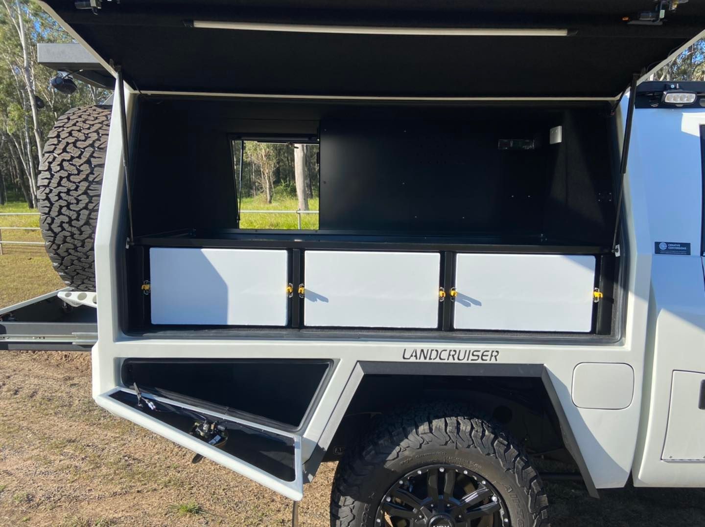 White truck bed with a canopy, storage drawers, and a spare tire. Black cargo rack mounted on a vehicle roof, with a protective grill at the rear. — Custom RV Creations & Repairs in Sumner, QLD
