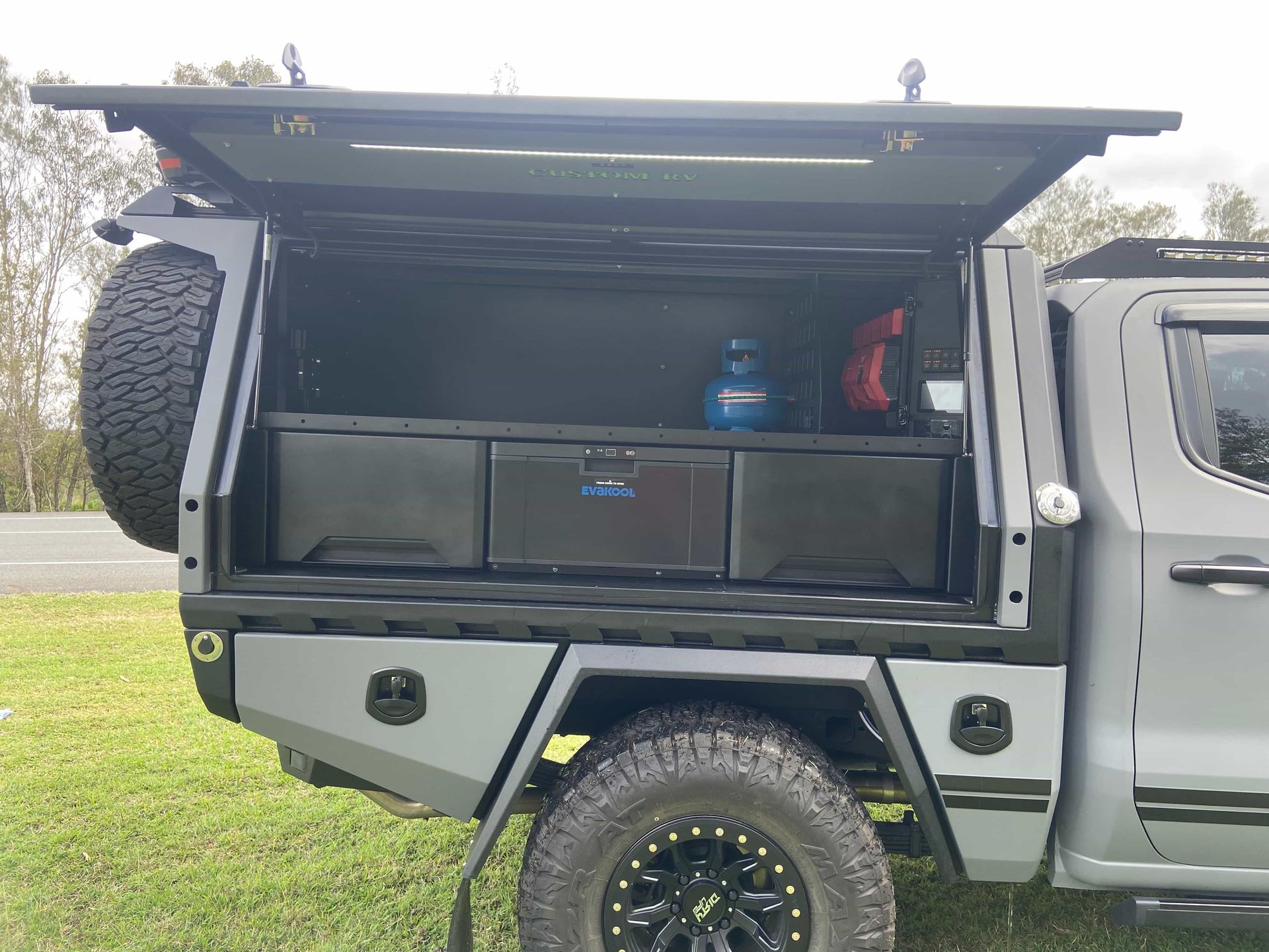 Gray truck bed with a storage unit, spare tire, and gear. Open door reveals drawers and a gas cylinder. — Custom RV Creations & Repairs in Sumner, QLD