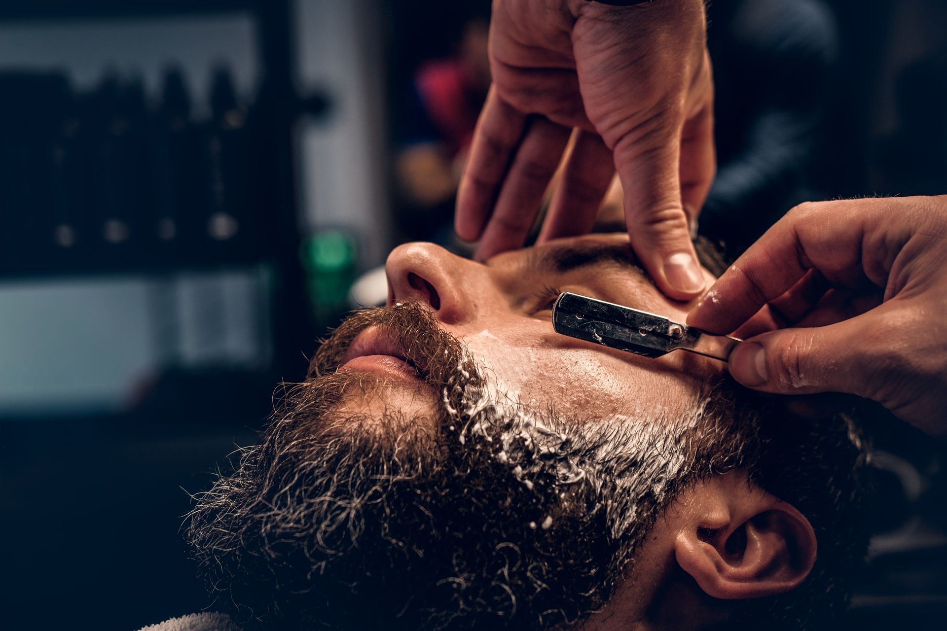 A person in a barber shop having their beard shaped with a straight razor while their skin is covered in shaving foam.