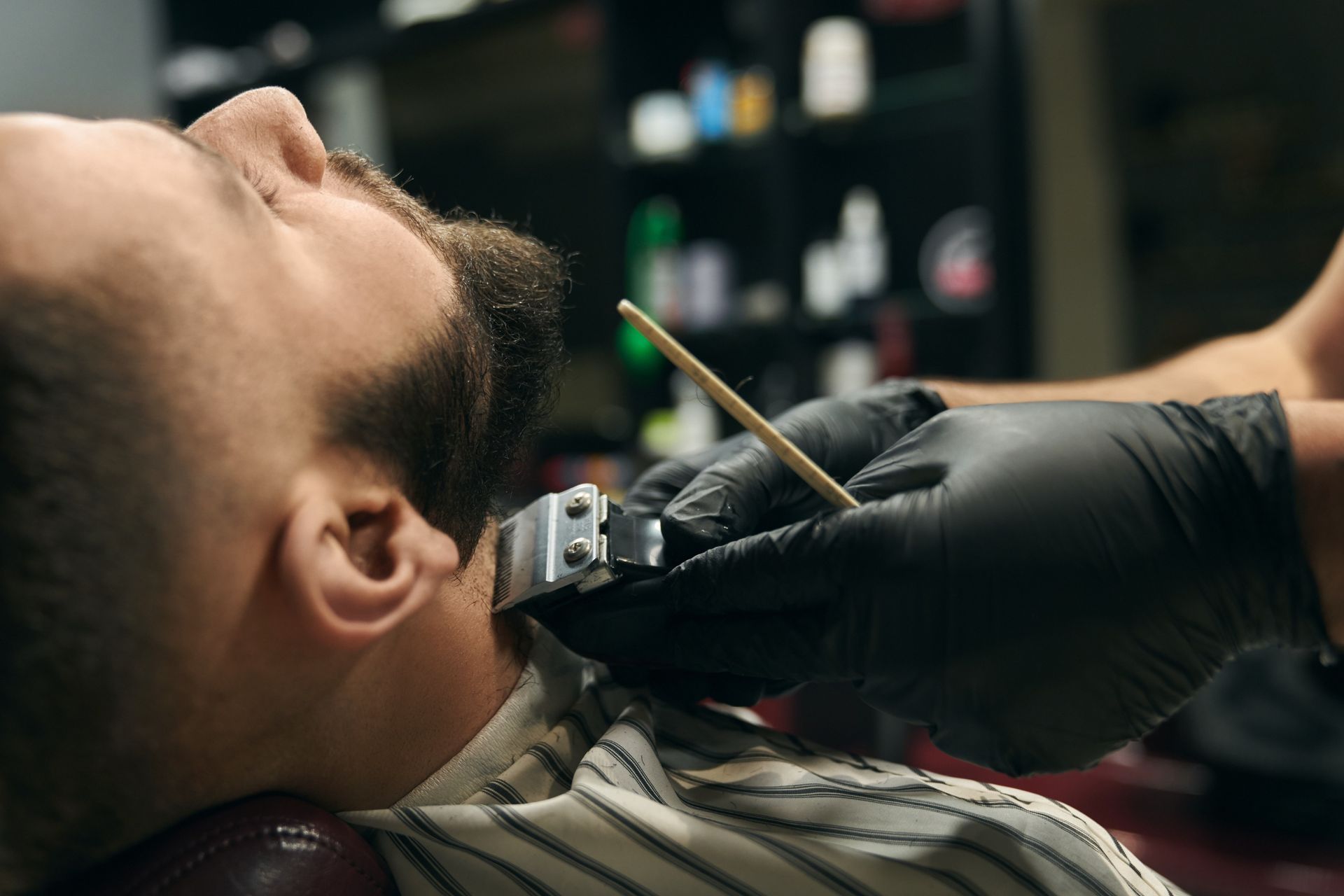 A barber wearing black gloves uses electric clippers to trim a person's beard in a salon.