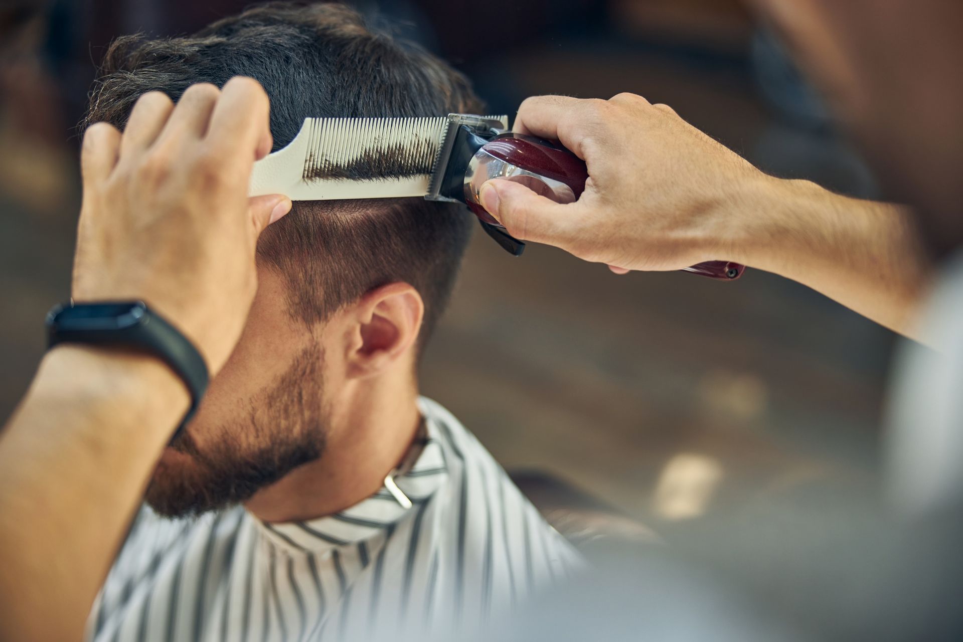 A barber uses clippers and a comb to trim hair on the side of a person’s head in a salon setting.