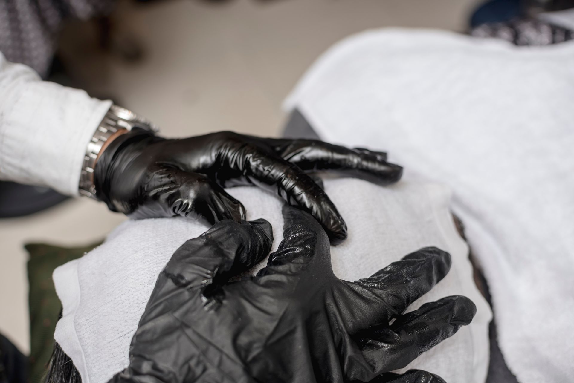 Two hands in black medical gloves gently touch a draped white cloth, suggesting a massage or spa treatment.