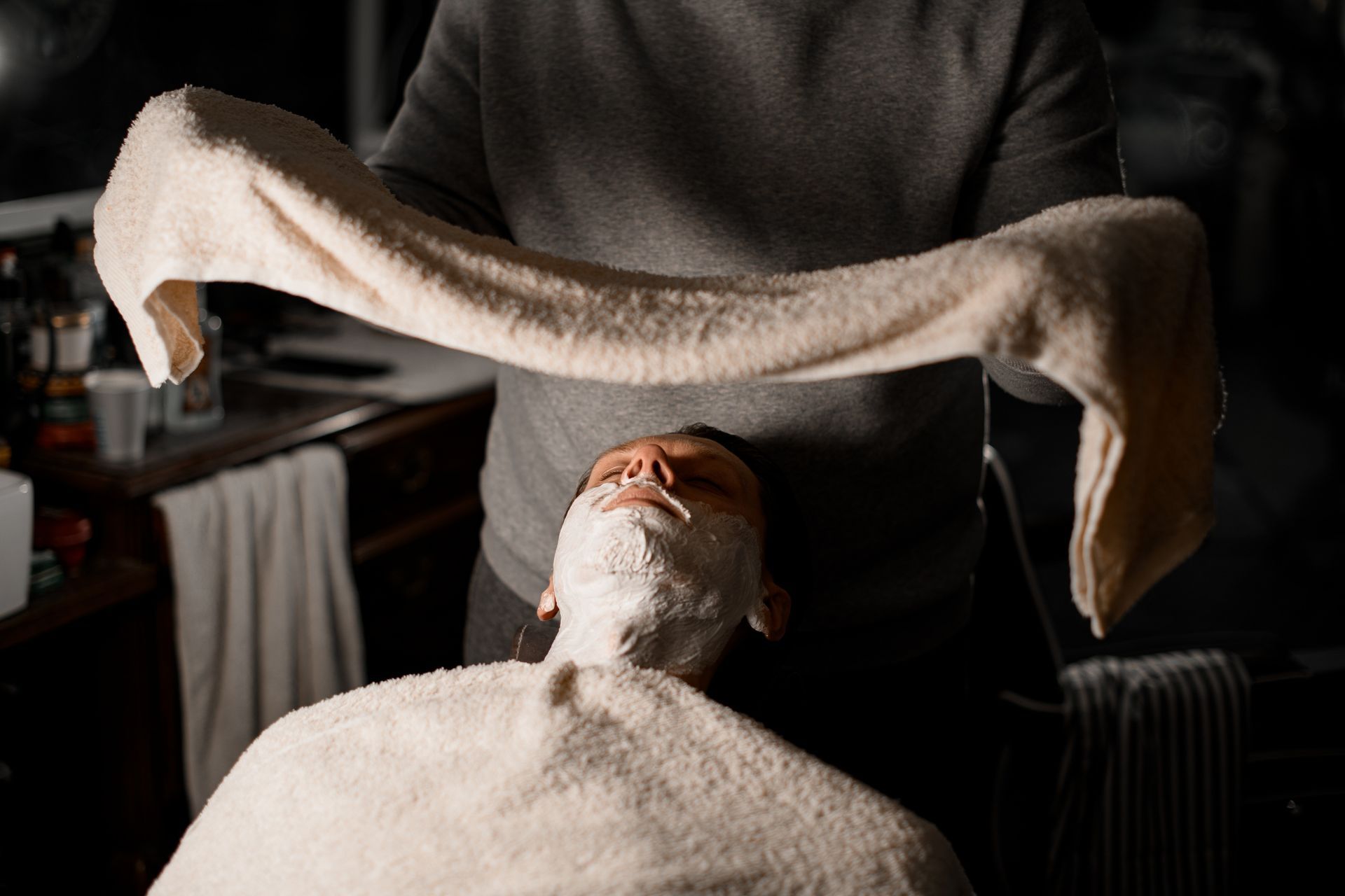 A barber holds a towel above a customer, who sits in a chair with white shaving cream covering their face.