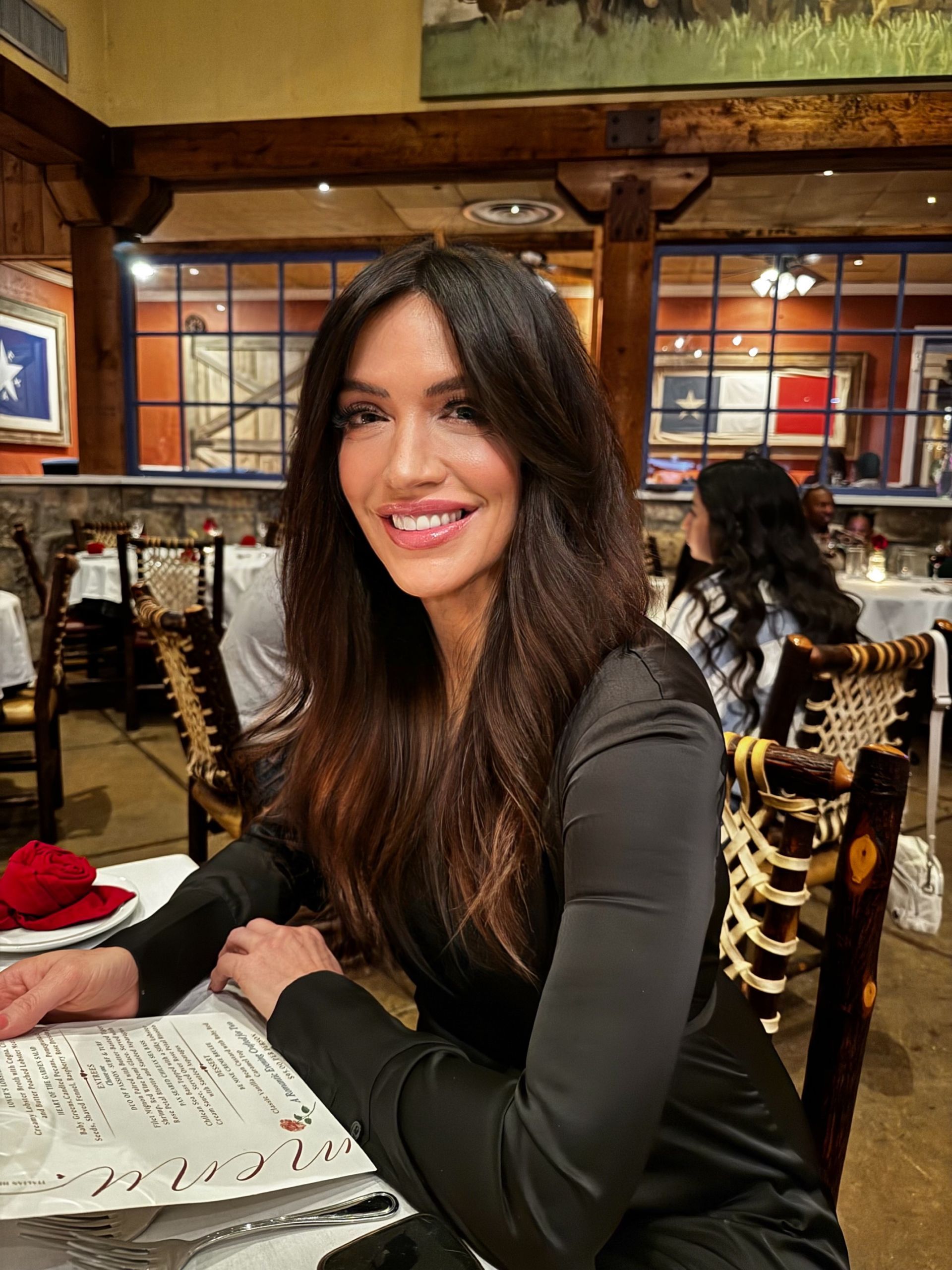 Woman with long dark hair smiling at a restaurant table, wearing a black top.