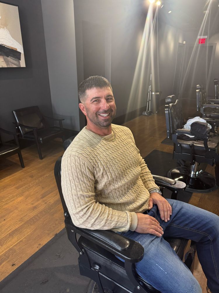 Man smiling in a beige sweater sitting in a salon chair under bright lights, with styling equipment nearby