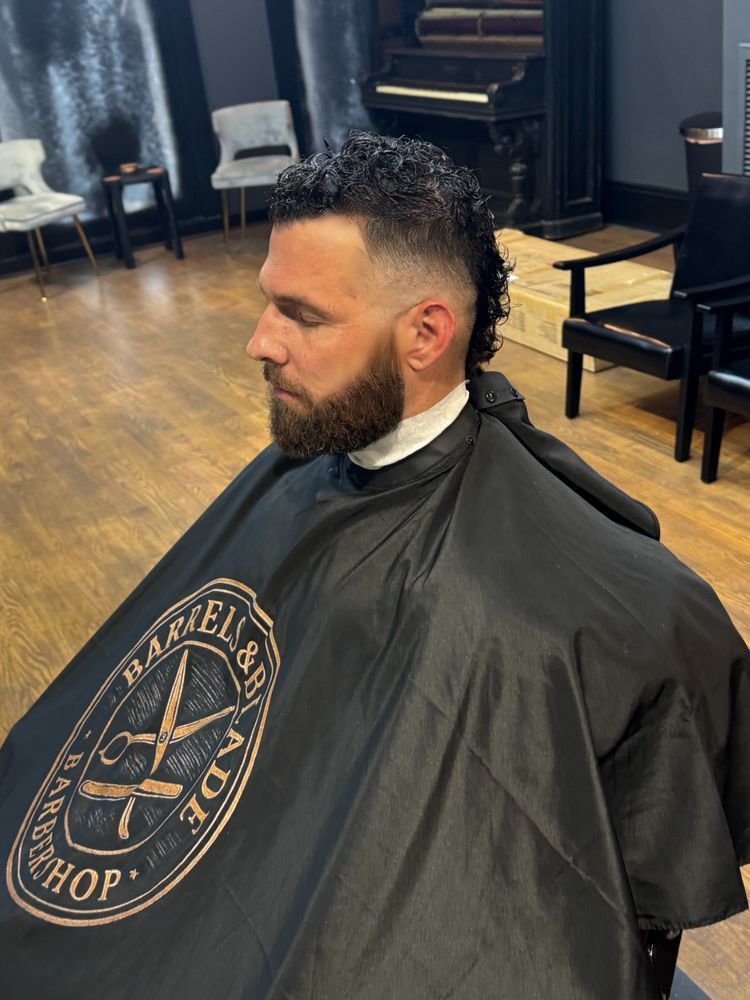 Man seated in a barber chair with a dark haircut cape on in a barbershop, viewed in profile.