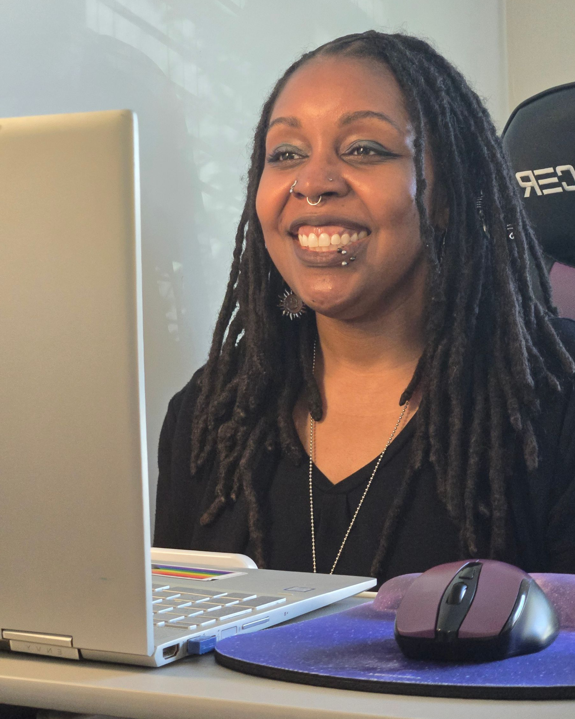 Woman with dreadlocks smiles at a laptop, sitting at a desk with a mouse.
