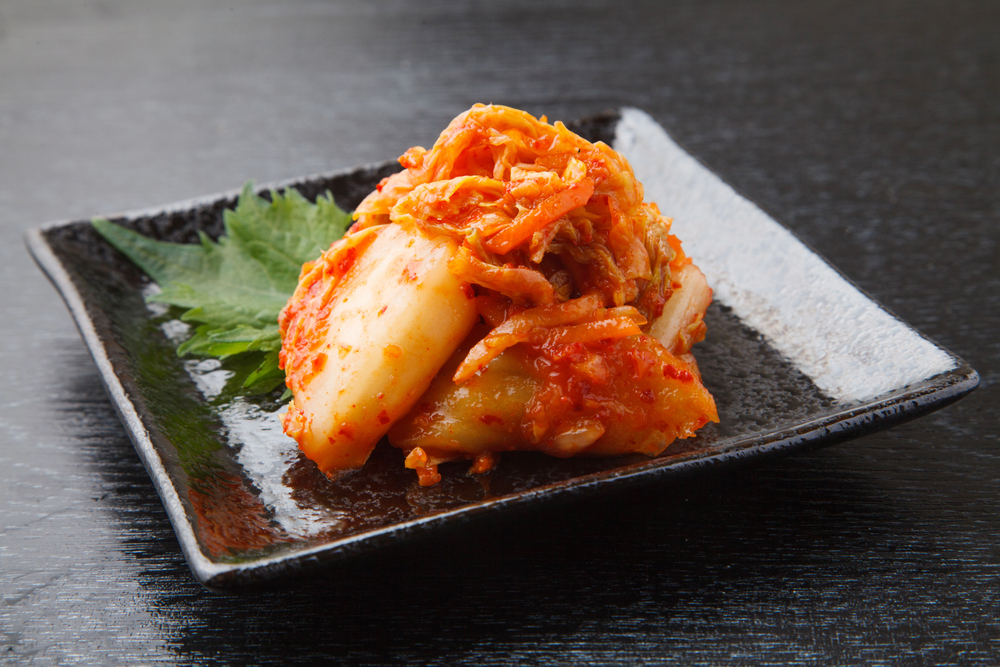 Kimchi on a small square plate with a green leaf. Red and orange fermented cabbage dish. Black background.